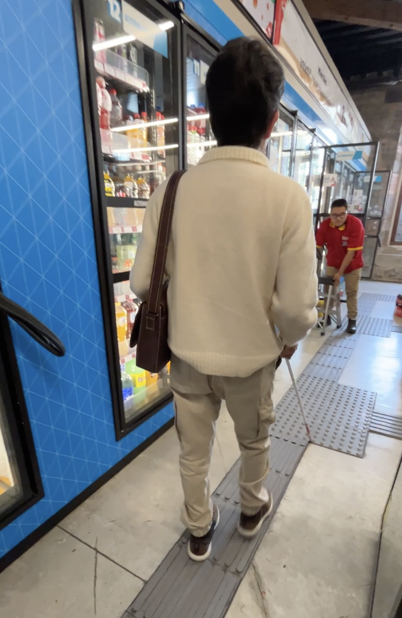 Foto de un hombre con discapacidad visual avanzando por la guía podotáctil que está por la tienda.